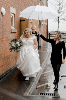 a bride walking in the rain being sheltered by somebody holding a white umbrella