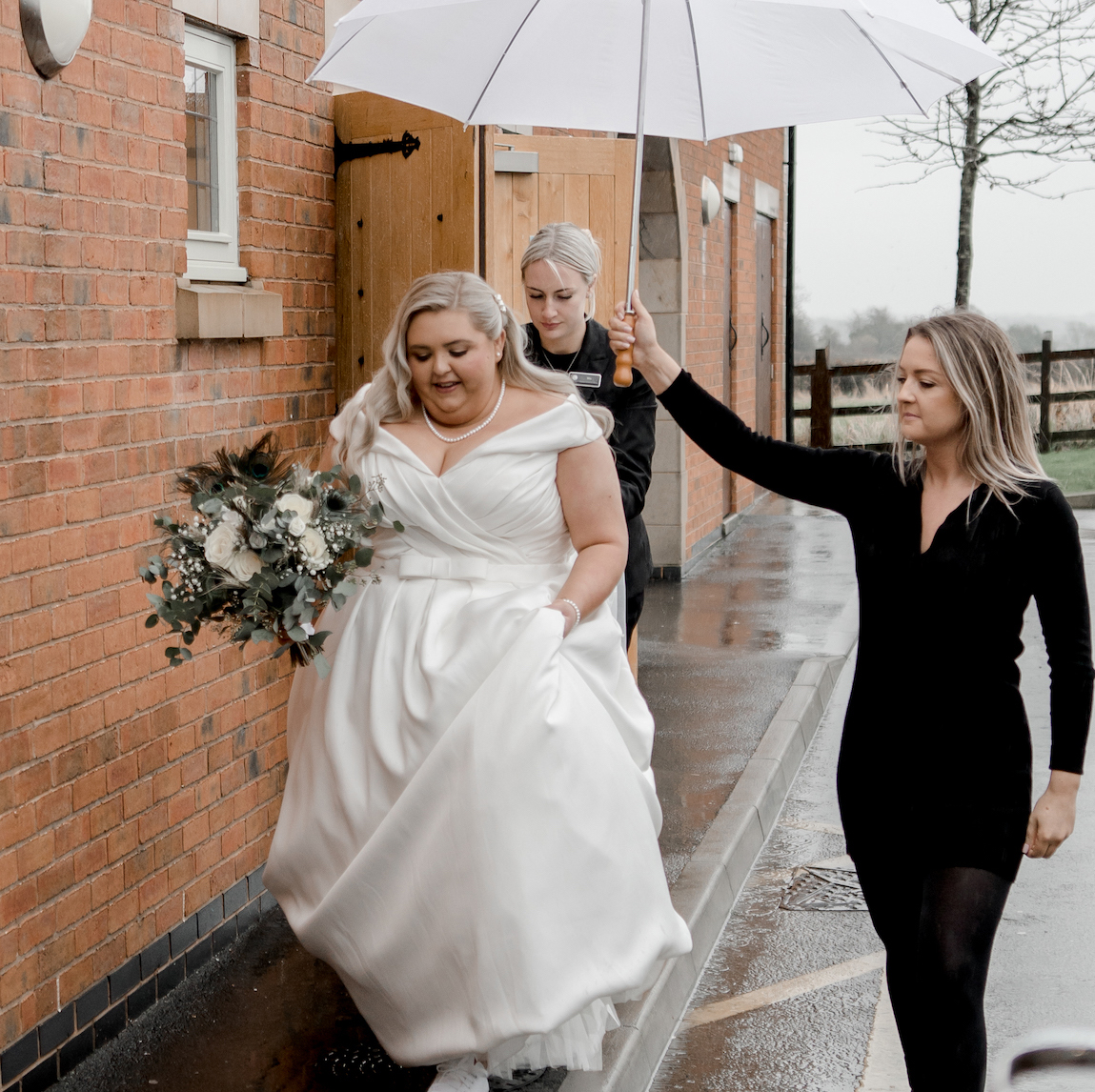 a bride walking in the rain being sheltered by somebody holding a white umbrella